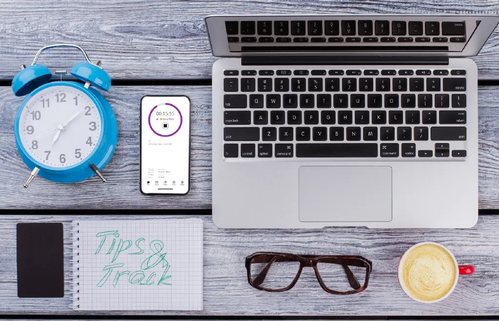 A desk with clock, laptop, glasses, notebook and smartphone with time tracking app